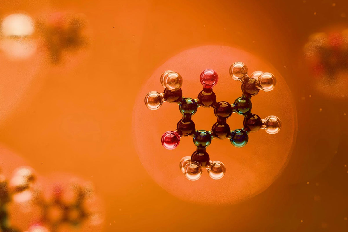 A close-up macro shot of a colorful molecular model of a caffeine molecule with red, green, and metallic silver spheres, set against a warm orange background with soft blurred bokeh.