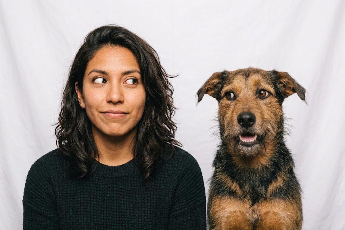 A woman with dark shoulder-length hair wearing a black knit top sits next to a brown and black dog against a white fabric backdrop. The woman and dog are side-eyeing each other.